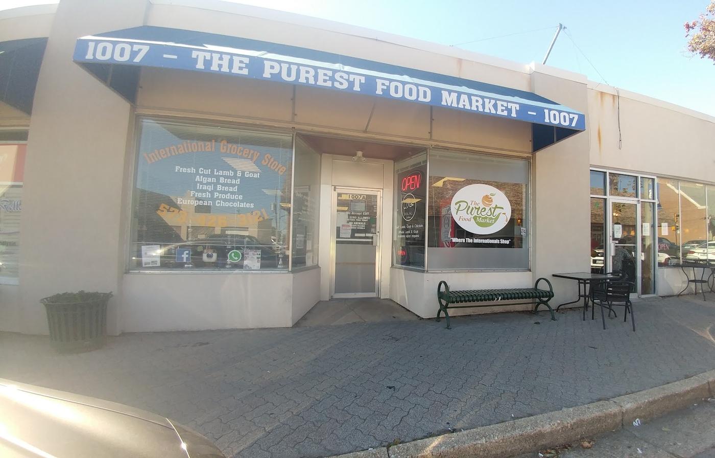 The storefront on North Pine Street: a blue awning reading 1007 The Purest Food Market, window lettering listing the goods, the round green and white logo on the glass, a neon open sign, and a bench and cafe table outside.